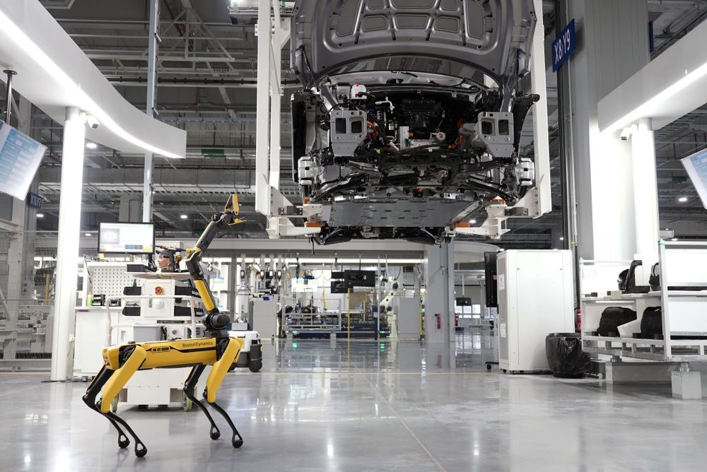A Boston Dynamics Spot quadruped with a manipulator arm walking a car factory floor.
