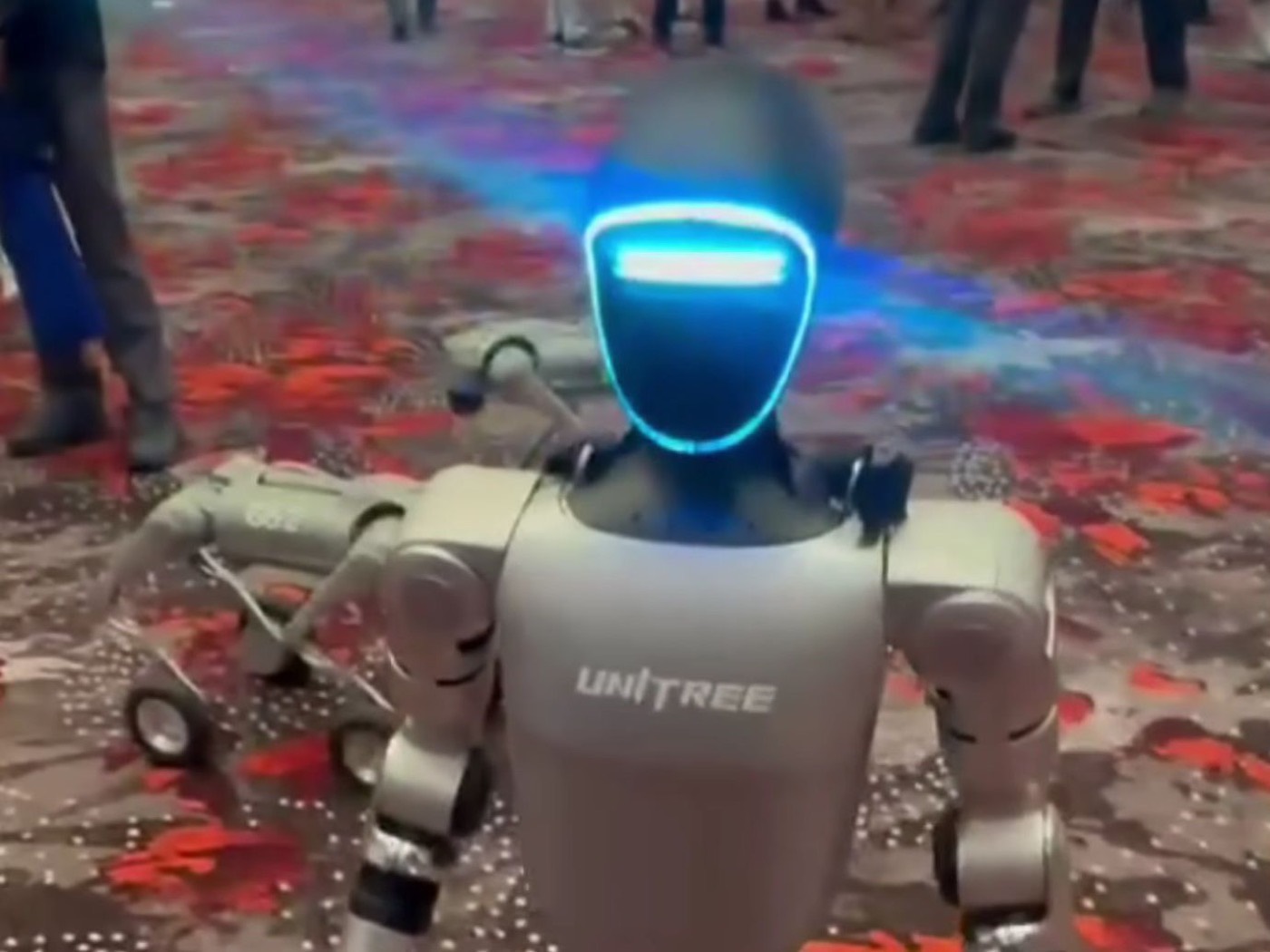 A Unitree G1 humanoid robot standing on an exhibition hall floor, its visor lit blue.